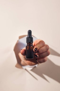 Bir kadının eli arka planda sert gölgeler olan yırtık bir kağıdın içinden bir şişe yüz yağını tutar. Güzellik kavramı.