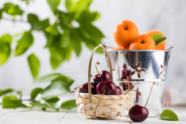 Dojrzałe wiśnie w koszyku i świeże morele w wiadrze na białym drewnianym tle. Naturalne produkty świeże. — Zdjęcie stockowe