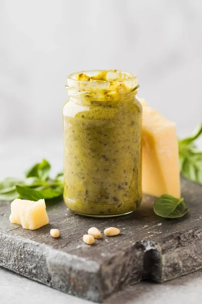 Salsa de pesto en un frasco con un trozo de queso parmesano, hojas de albahaca, piñones en una tabla de madera. Cocina italiana — Foto de Stock