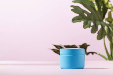 Krema için mavi bir kavanoz, yeşil bitkili pembe bir arka planda kozmetik maske. Boşluğu kopyala.