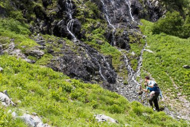 Sırt çantasında oturan bir baba ve bir çocuk dağ şelalelerinde duruyorlar. Çocuklarla aktif eğlence.