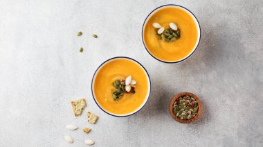 Gri zemin üzerinde balkabağı kremalı çorba ve kabak çekirdekli iki kase. Üst Manzara. Sonbahar tarifleri.