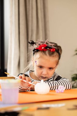 A cute girl paints a Halloween pumpkin with a headband for a festive craft activity. Concept idea for a kids craft.