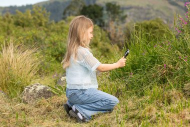 Young caucasian girl exploring nature with magnifying glass in mountainous landscape. An inquisitive girl explores nature.