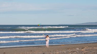 Güneşli bir günde renkli bir uçurtma uçururken sahilde koşan mutlu küçük kız. Çocuk deniz kenarında eğleniyor ve özgürlüğün tadını çıkarıyor. Aile tatili sırasında eğlenceli ve kaygısız çocukluk anı.