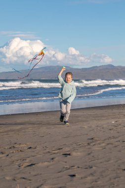 Neşeli genç bir çocuk kumsalda koşuyor ve rüzgarlı bir günde renkli bir uçurtma uçuruyor. Deniz kenarındaki çocuklar için eğlenceli bir açık hava aktivitesi