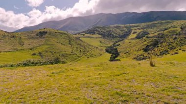 Mavi gökyüzü ve bulutların altında yuvarlanan yeşil tepeleri olan görkemli dağ manzarası. Yeşil tepeler ve mavi gökyüzü ile Yeni Zelanda dağ manzarası, eko-turizmi ve sürdürülebilir yolculuğu sembolize ediyor.