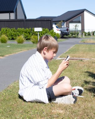 Genç bir çocuk, güneşli bir günde yeşil çimenlerde akıllı telefon kullanarak oturuyor, dışarıda geziniyor ya da mesaj yazıyor. Teknoloji ve doğa ile rahat çocukluk anı.