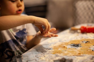 Mutfak masasında hamur hazırlarken elleri un kaplı, evde kurabiye pişiren küçük bir çocuk. Sıcak ortam aydınlatması sıcak bir aile ortamı yaratır.