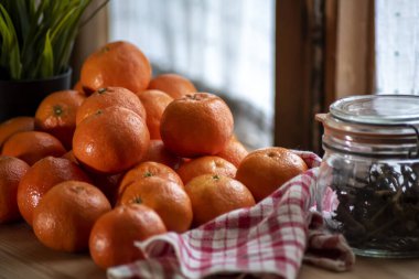 Mandalinalar (portakallar, mandalinalar, narenciye) cam kavanoz ve kareli peçete ile ahşap masa üzerinde, perde ve arka planda bitki olan pencere. Seçici odak