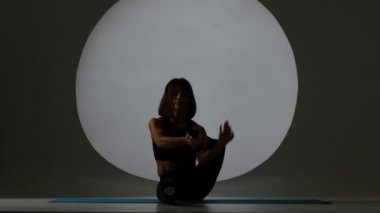 Jimnastikçi mindere oturmuş bir bacak arkasında kafasını alır. Arka ışık. Siluet