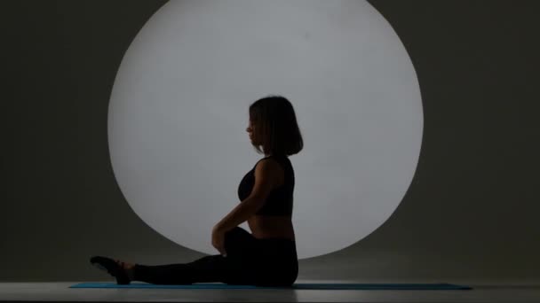 Kvinna sitter på yogamattan och stretching benen. Bakgrundsbelysningen. Siluett — Stockvideo