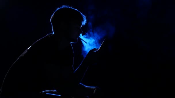 Мужчина смотрит на телефон и курит сигарету. Свет сзади. Силуэт — стоковое видео