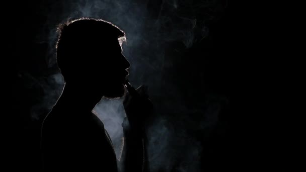 El hombre fuma cigarrillos electrónicos. Negro. Luz trasera. Silueta. Movimiento lento — Vídeo de stock