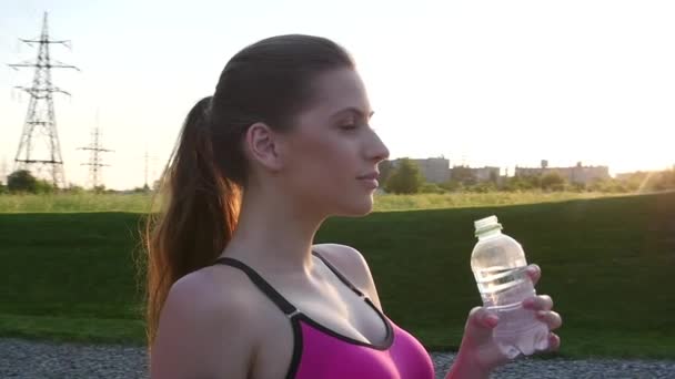 Fitness kvinna dricksvatten utomhus i parken mot gyllene solnedgången himmel. Slow motion — Stockvideo