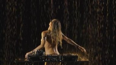 Vücudunda gümüş bir vücut boyası olan çıplak bir disk jokeyi yağmurda kumanda üzerinde çalışır. Gece hayatı, parti konsepti. Kapatın. Yavaş çekim.