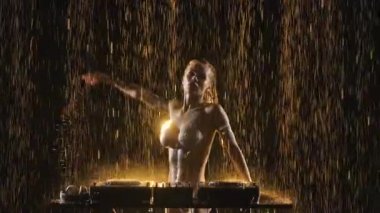 Seksi sarışın dans eder ve DJ konsolunda yağmur damlaları ve stüdyo ışıkları arasında müzik karıştırır. Kadınlar saçlarını dalgalandırır ve müzikten zevk alırlar. Disko ve moda etkinlikleri. Kapatın. Yavaş çekim.