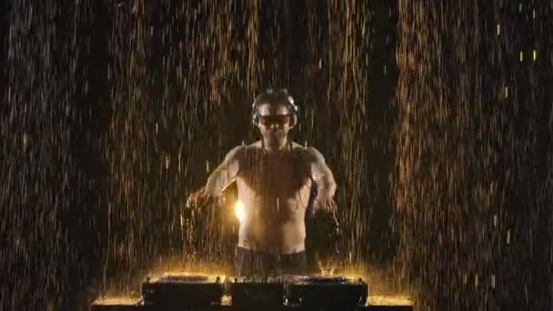 一个很酷的留胡子DJ站在一个搅拌机控制器后面，创作了一个新的混音，并在雨中的黑暗工作室里点燃了一个派对。DJ带着眼镜和耳机创作舞曲。靠近点慢动作. — 图库视频影像