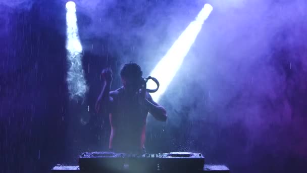 Silhouette of male DJ with headphones and in sunglasses dances and mixes music with a mixer controller in a dark smoky studio with blue light. Disco in the rain at night. Close up. Slow motion. — Stock Video