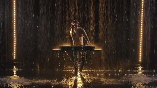 Un DJ barbu dans les tatouages joue de la musique sur un contrôleur de mixeur sous la pluie dans un studio sombre. Les ruisseaux d'eau sont magnifiquement éclairés de lumière jaune. Mouvement lent. — Video