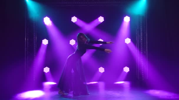 Genadige jonge vrouw in zwart lichaam danst contemp in studio duisternis. De slanke actrice steekt haar armen soepel op in een sensuele dansbeweging tegen de paarse achtergrondverlichting in de rook. Langzame beweging. — Stockvideo
