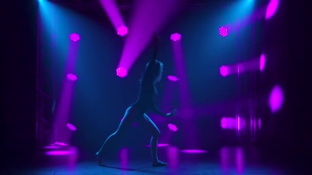 Silhouette d'une fille gracieuse pratiquant le mépris dans l'obscurité du studio. Elle danse des éléments de danse sur un fond noir avec des projecteurs colorés dynamiques. Mouvement lent. — Video