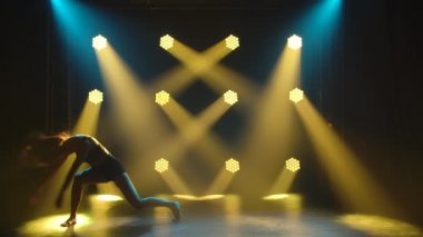 Siyah iç çamaşırlı zarif bir kadının, sarı spot ışıklarının önünde stüdyonun karanlığında aşağılama alıştırması yapması. Siluet. Yavaş çekim.