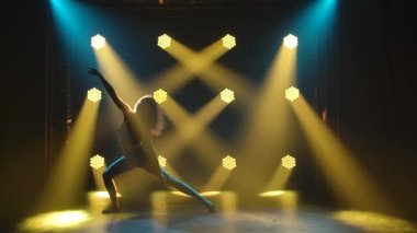 Siyah iç çamaşırlı zarif bir kadının, sarı spot ışıklarının önünde stüdyonun karanlığında aşağılama alıştırması yapması. Siluet. Yavaş çekim.