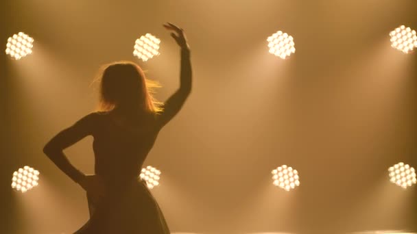 Silhouet van een jonge mooie vrouw in een zwart bodysuit en rok die een moderne hedendaagse balletdans danst. Close-up genomen in een donkere studio met geel licht. Langzame beweging. — Stockvideo