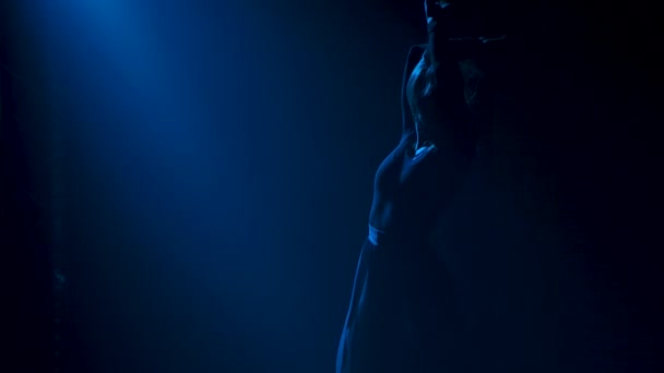 Тонкий силуэт привлекательной танцовщицы, исполняющей эмоционально современную хореографию. Молодая женщина танцует в тёмной дымчатой студии на фоне синих огней. Закрывай. Медленное движение. — стоковое видео