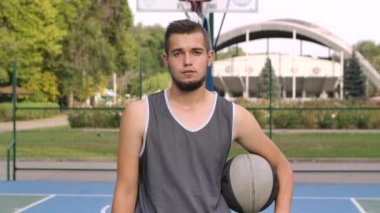 Yakışıklı sakallı beyaz basketbolcunun portresi oyun alanında kameraya bakıyor. Sporcu, elinde topla açık havada sahada duruyor. Kapatın. Yavaş çekim.