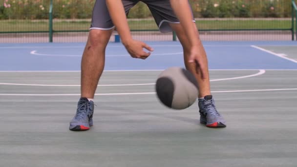 Een basketbalspeler dribbelt tussen z 'n benen op het veld. Een atleet in een grijs sportuniform slaat de bal op de grond en kijkt naar de camera. Benen dicht. Langzame beweging. — Stockvideo