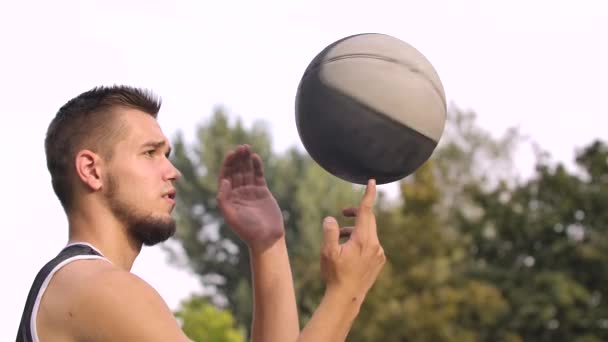 Ung basketspelare står på planen utomhus och snurrar boll på fingret. Man i sportkläder vilar efter streetball match på gatan. Idrottsman tränar färdigheter. Närbild. Långsamma rörelser. — Stockvideo
