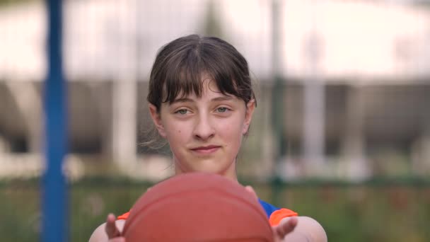 Porträt eines jungen Mädchens, das in die Kamera blickt und einen Basketball nach vorne reckt. Ein Teenager in blau-oranger Sportuniform bereitet sich auf ein Basketballspiel vor. Aus nächster Nähe. Zeitlupe. — Stockvideo