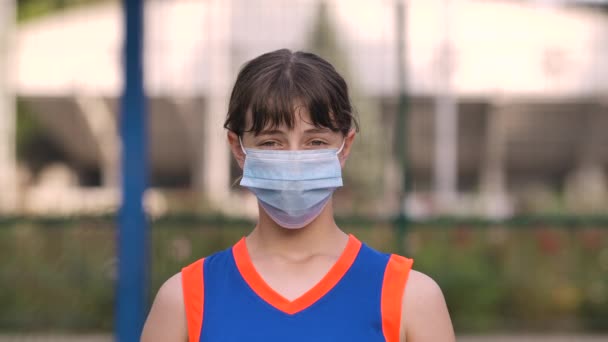 Kameraya bakan tıbbi maskeli genç bir kız basketbolcunun portresi. Koronavirüs karantinası sırasında basketbol maçına hazırlanan bir genç. Kapatın. Yavaş çekim. — Stok video