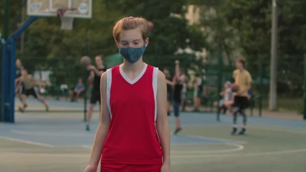 Egy védőmaszkos lány portréja, aki a városi játszótéren edz egy kosárlabdával. Egy piros sportegyenruhás tinédzser sportolni megy a Covid 19 karantén alatt. Közelről. Lassú mozgás. — Stock videók