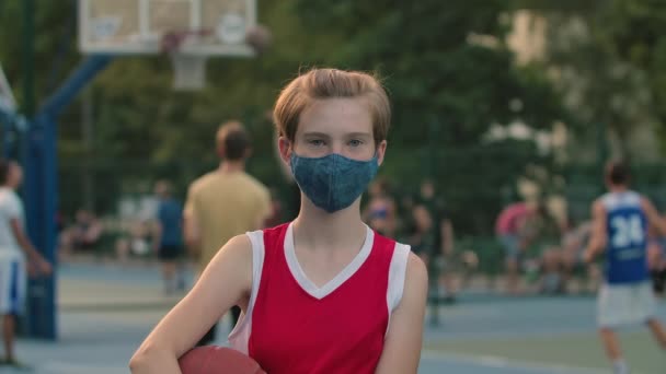 Egy védőmaszkos lány portréja, aki egy városi játszótéren áll egy kosárlabdával. Egy piros sportegyenruhás tinédzser készül egy játékra a Covid 19 karantén alatt. Közelről. Lassú mozgás.. — Stock videók