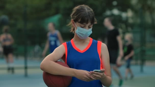 Mladá dívka v lékařské masce s míčem píše textovou zprávu na svém smartphonu. Zavřít. Zpomalený pohyb. — Stock video