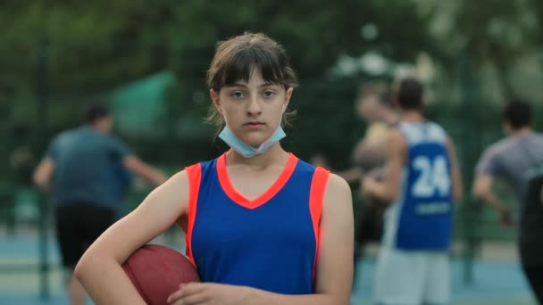 Dziewczyna koszykarz z piłką patrzy w kamerę i zakłada maskę medyczną. Sporty na świeżym powietrzu i ochrona przed koronawirusem. Zamknij drzwi. Zwolniony ruch. — Wideo stockowe