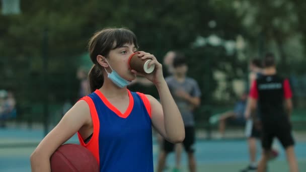 Une adolescente portant un masque médical abaissé se tient sur un terrain de basket-ball de sport de rue avec une balle buvant du café et se réjouissant. Ferme là. Mouvement lent. — Video