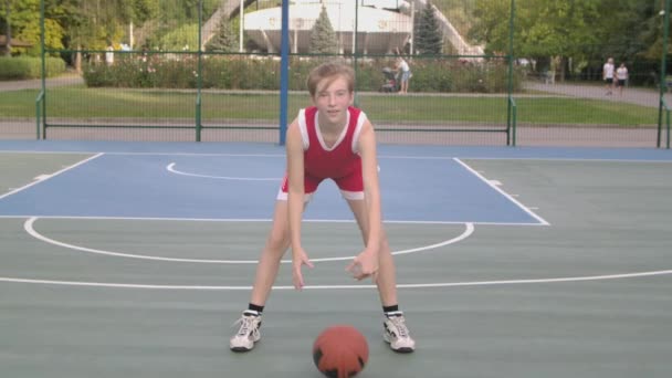 Ung flicka spelar basket utomhus öva bollhantering skicklighet. En lång tjej basketspelare i röd uniform står på lekplatsen och ser straight ut. Närbild. Långsamma rörelser. — Stockvideo