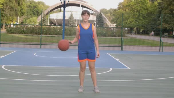 Retrato de una joven jugador de baloncesto está de pie con la pelota y mirando a la cámara. Un adolescente en uniforme deportivo naranja azul preparándose para un partido de baloncesto. De cerca. Movimiento lento. — Vídeo de stock