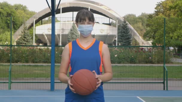 Portret młodej koszykarki w masce medycznej patrzącej w kamerę. Nastolatek przygotowujący się do gry w kosza podczas kwarantanny. Zamknij drzwi. Zwolniony ruch. — Wideo stockowe