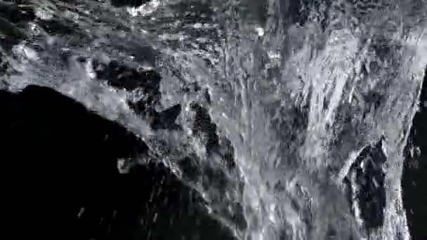 Ett objekt faller på ytan uppifrån och stänk vatten över en svart bakgrund. Bakgrundsbelysta stänk glittrar i studioljus. Explosionsvatten i slow motion närbild. — Stockvideo
