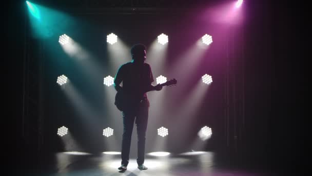 Le folklore africain. Un homme pieds nus joue de la guitare dans un studio sombre sur fond de lumières vives. Des silhouettes. Mouvement lent. — Video