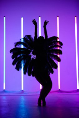 Brezilya karnaval kostümünde tüylerden yapılmış ince bir dansçının silueti. Kadın stüdyoda arka planda mor ışıklandırmalı neon lambalara karşı poz veriyor..
