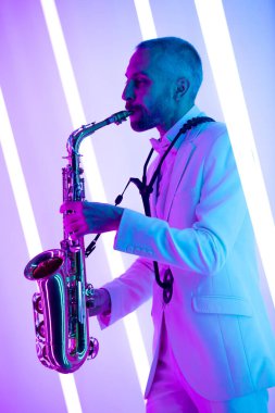 Stüdyoda beyaz takım elbiseli, şık bir saksafoncunun portresi. Profesyonel bir müzisyen neon lambaların arka planına karşı saksafon çalar. Resmin dikey yönelimi.