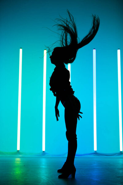 Side view of a young slender woman with flying hair posing standing in the studio against a background of neon lamps with blue backlight