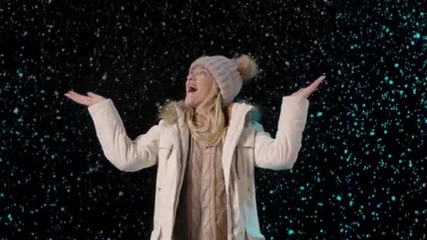 Portret van een jonge vrouw die geniet van de sneeuw. Een blondine in warme winterkleding op een zwarte achtergrond verheugt zich bij de eerste sneeuw. Fluffy grote sneeuwvlokken vallen rond. Langzame beweging. Sluiten.. — Stockvideo