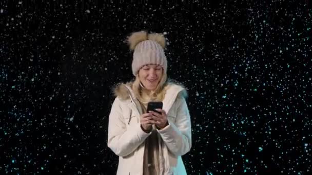 Retrato de uma bela jovem escreve mensagens de texto em seu telefone enquanto está em pé entre a neve caindo. Loira alegre em roupas quentes de inverno em um fundo preto. Movimento lento. Fechar. — Vídeo de Stock